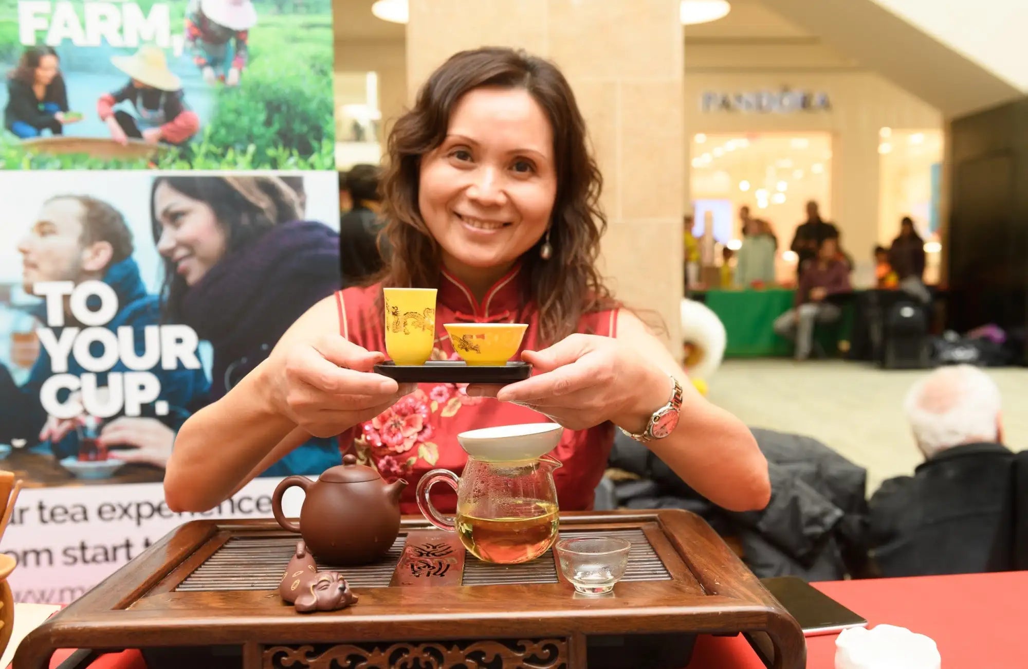 Yellow ceramic tea cups with delicate floral patterns, held on a black tray by a smiling woman in a red dress.