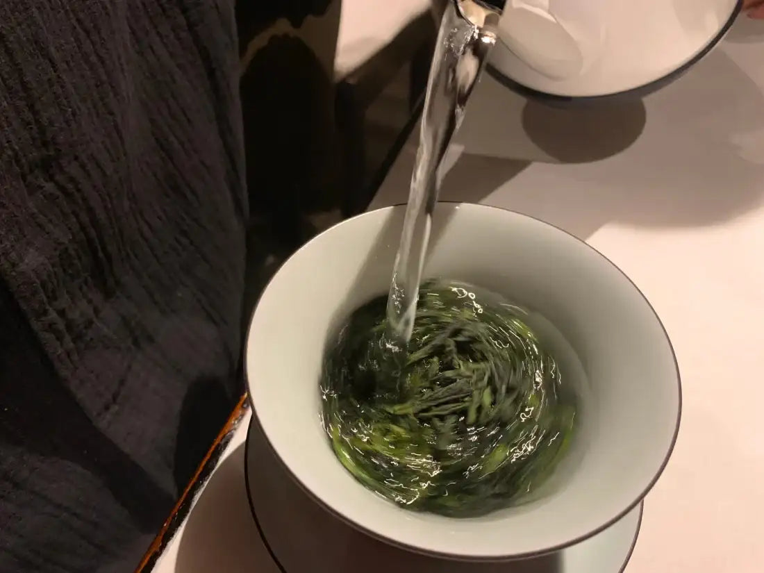 A white ceramic teacup filled with swirling green tea leaves and clear water, being poured from a matching pitcher.