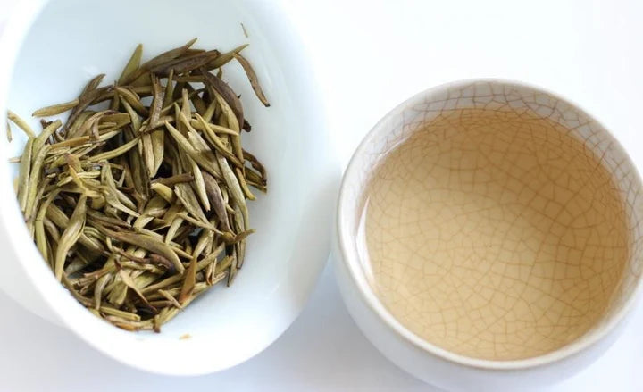 A delicate, pale yellow-green tea leaves in a white ceramic bowl beside a matching crackled-glazed teacup filled with brewed tea.