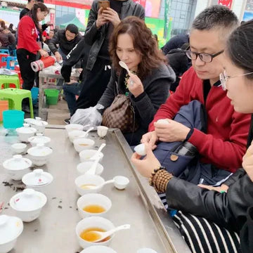A long, metallic table holds a row of white ceramic tea bowls and spoons filled with golden liquid.