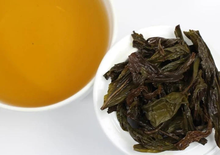 A cup of golden amber tea beside a white bowl filled with dark, twisted oolong leaves.