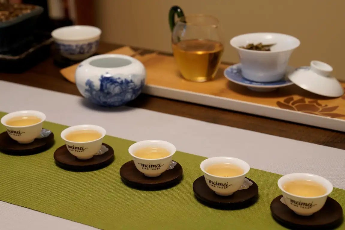 A row of five white ceramic tea cups with ’meimei’ branding, each resting on a dark brown circular coaster.